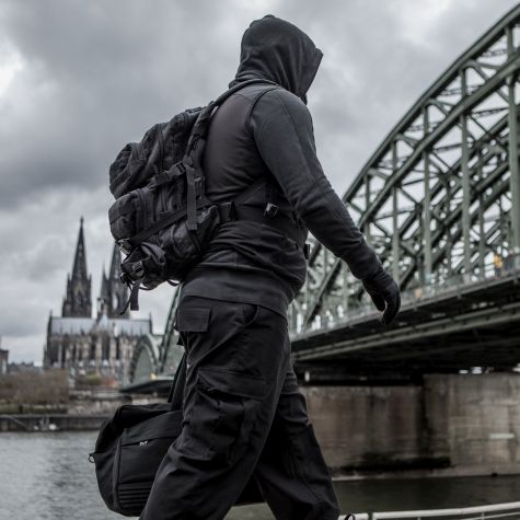Die Aufnahme zeigt einen einfarbig angezogenen Mann mit Kapuzenpullover, der unerkannt bleiben möchte und an einem Flussufer eine Reisetasche trägt