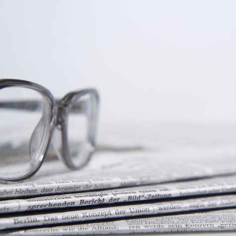 Die Aufnahme zeigt einen Stapel Zeitungen vor einem weißen Hintergrund, auf dem eine Brille liegt.