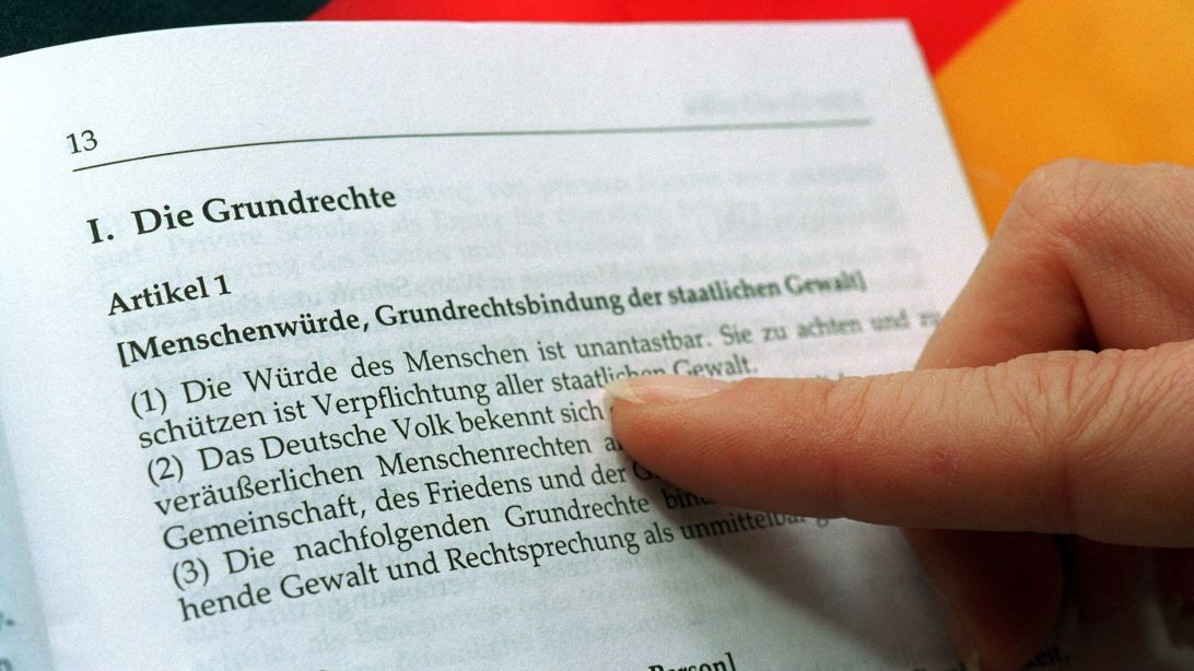 The photograph, taken on April 19, 1999, shows a female index finger pointing to Article 1 of an open edition of the Basic Law. A German flag can be seen in the background. (Source: Stephanie Pilick/dpa/picture alliance) The photograph, taken on April 19, 1999, shows a female index finger pointing to Article 1 of an open edition of the Basic Law. A German flag can be seen in the background.